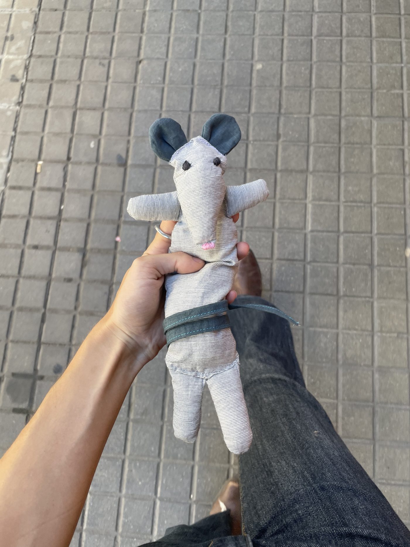 A hand-sewn fabric mouse in grey striped cotton, with dark ears and a pink stitched nose, held up in one hand above a tiled pavement.